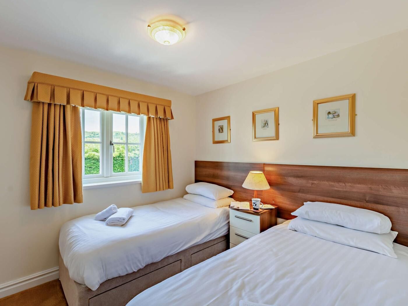 A hotel room with two beds, a window with curtains, a bedside lamp, and three framed pictures on the wall.