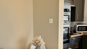 Et spiseområde med et rundt bord, hvide stole og en vase med tørrede blomster. Et køkken med mørke skabe og hårde hvidevarer er synligt i baggrunden.