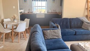 En moderne stue med en blå sofa, et spisebord med stole, og et køkken der kan ses igennem en åben døråbning.