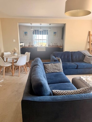Ein modernes Wohnzimmer mit einem blauen Sofa, einem Esstisch mit Stühlen und einer durch einen offenen Durchgang einsehbaren Küche.