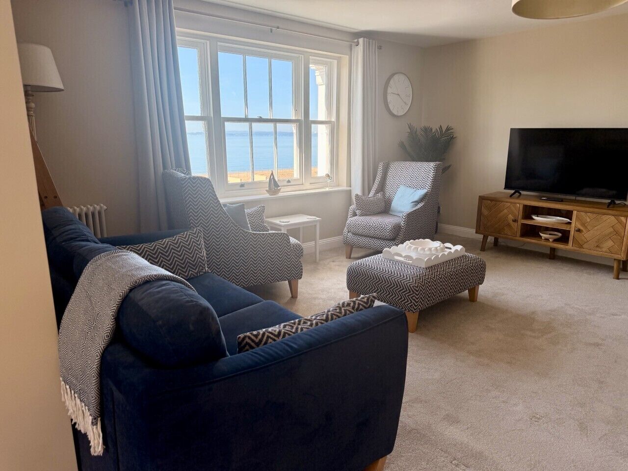 A living room with a blue sofa, two armchairs, a small table, a television, and a view of the sea through a large window.