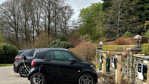 Um carro preto estacionado em uma estação de carregamento em uma área ajardinada com muros de pedra e árvores.