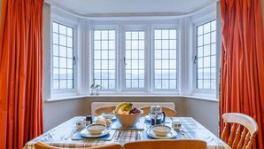 Uma sala de jantar com uma mesa posta para quatro pessoas, janelas grandes com cortinas e vista para o exterior.