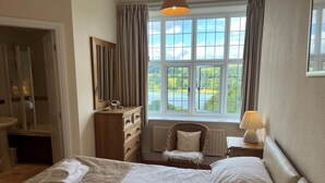 Ein Schlafzimmer mit einem Bett, einem Schminktisch, einem Sessel, einer Lampe und einem Fenster mit Blick auf Grünflächen.