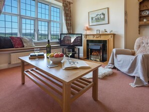 A cozy living room with a fireplace, a large window with a view, a comfortable sofa, and a table with a bowl of fruit and glasses. - 2 Bed in Hawkshead (oc-llh32) (Claife)
