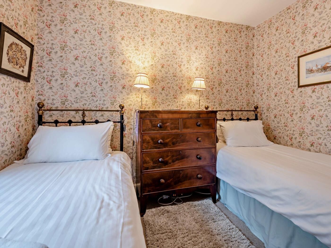 Une chambre avec deux lits simples, une commode en bois, un papier peint à motifs floraux et deux lampes fixées au mur.