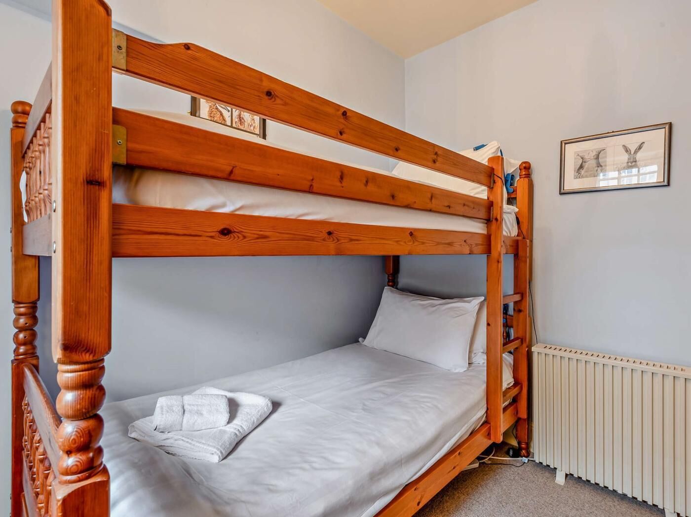 A bunk bed with wooden frames and white bedding, a radiator, and a framed picture on the wall.