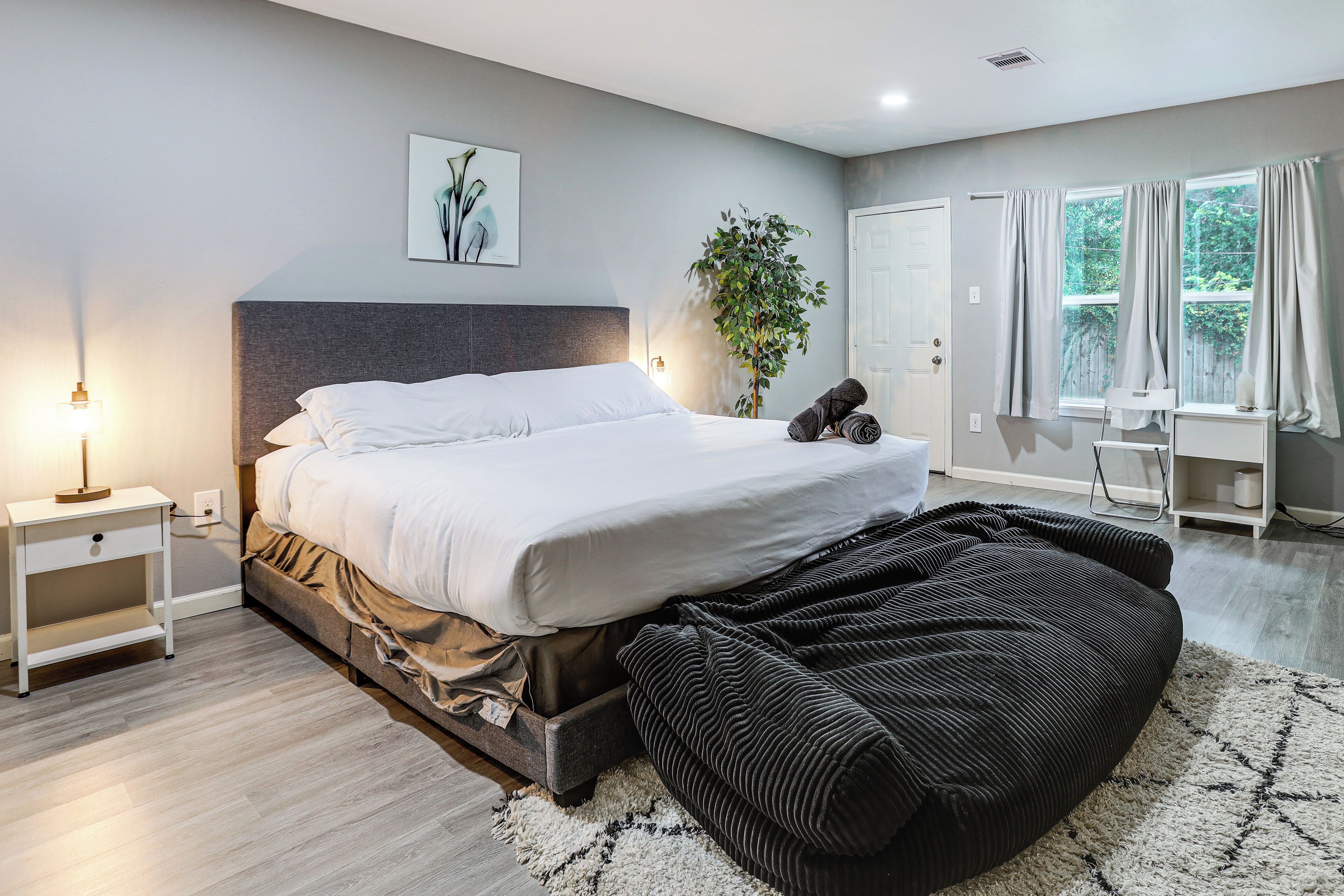 Une chambre à coucher moderne, avec un lit, une table de chevet, une lampe, une plante et une vue sur l’extérieur par une fenêtre.