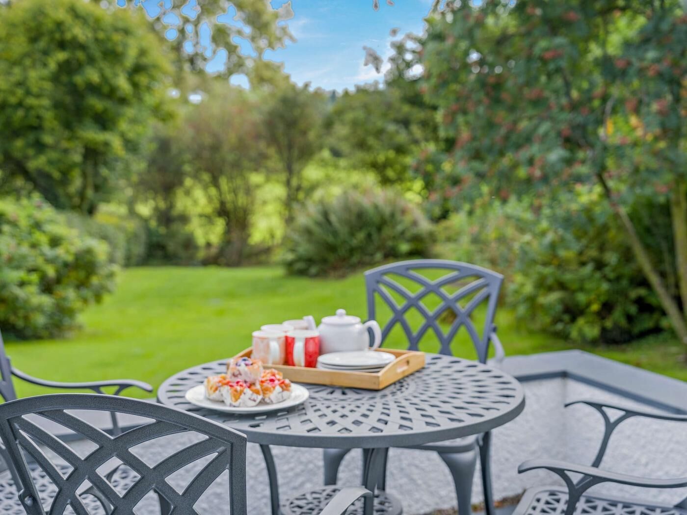 A round metal table with chairs, a tray with food, and a teapot in a garden setting.