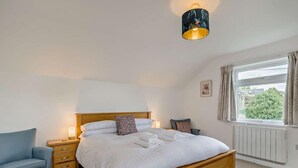 Ein Schlafzimmer mit einem hölzernen Bett, Nachttischen, einem Sessel, einem Fenster mit Vorhängen und einer Wandlampe.