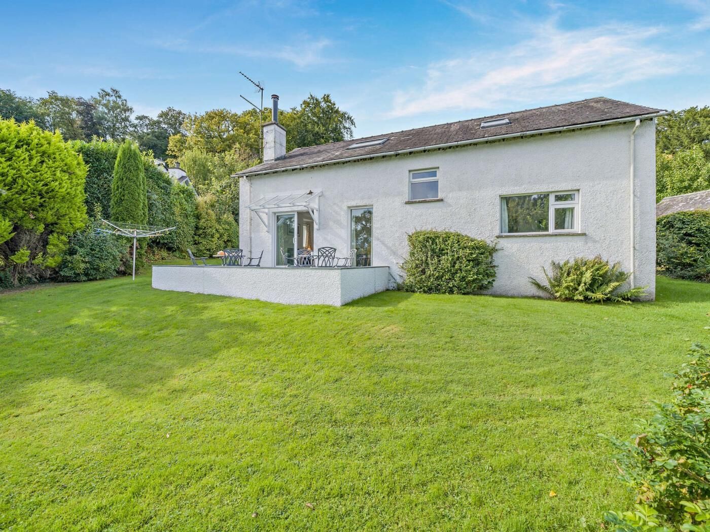 Ein eingeschossiges Haus mit weißer Fassade, einer kleinen Veranda mit Stühlen und einem gepflegten Rasen.