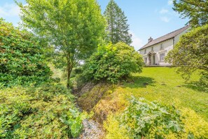 A lush garden with a variety of plants and trees, a small stream, and a traditional house with a white facade and dark roof. - 4 Bed in Hawkshead (oc-llh44) (Claife)