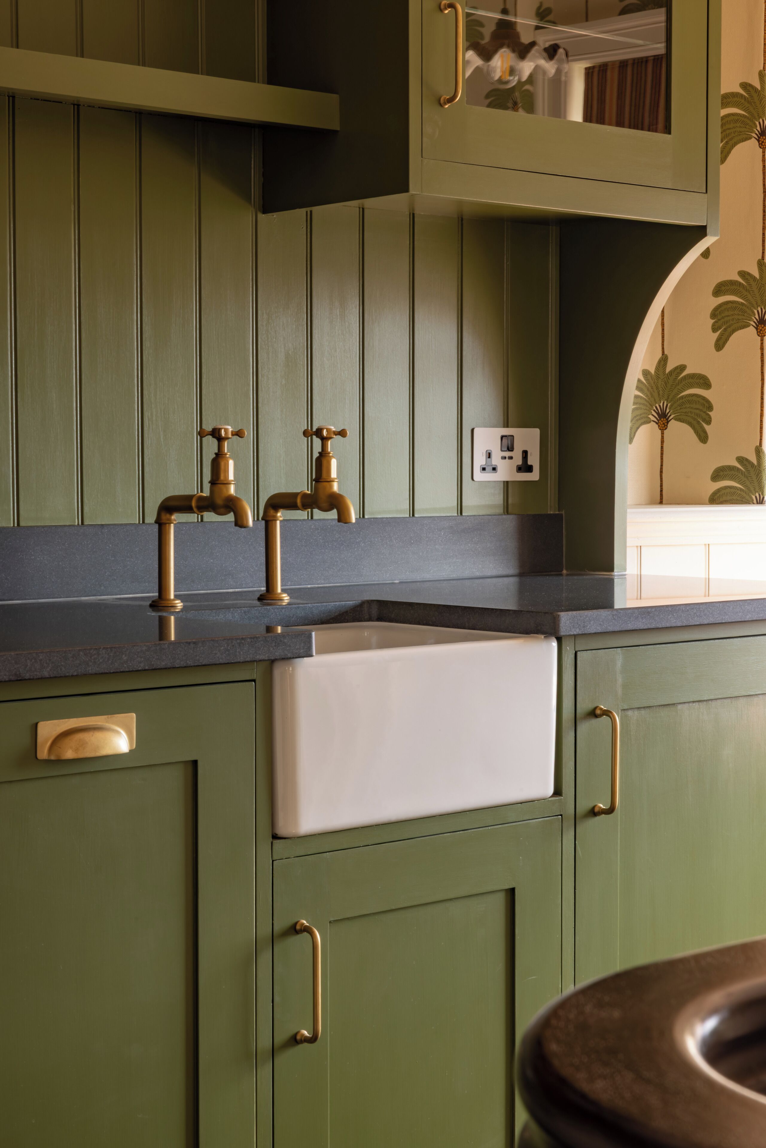 A kitchen with green cabinets, a white sink, and brass faucets.