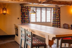 A kitchen with a central island, wooden chairs, a hanging light fixture, and a window with patterned curtains.