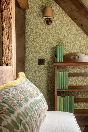 A room with a patterned wallpaper, a bed with a colorful blanket, a lamp, and a bookshelf with green books.