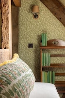 A room with a patterned wallpaper, a bed with a colorful blanket, a lamp, and a bookshelf with green books.