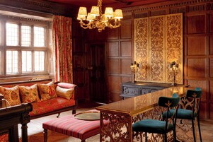 A room with a wooden paneled wall, a large window with curtains, a sofa with patterned cushions, a round table with chairs, and a chandelier.