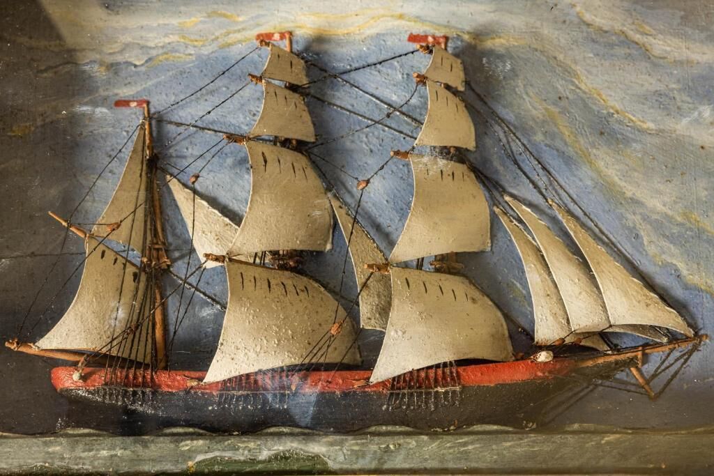 A model ship with multiple sails and a red hull, displayed against a textured background.