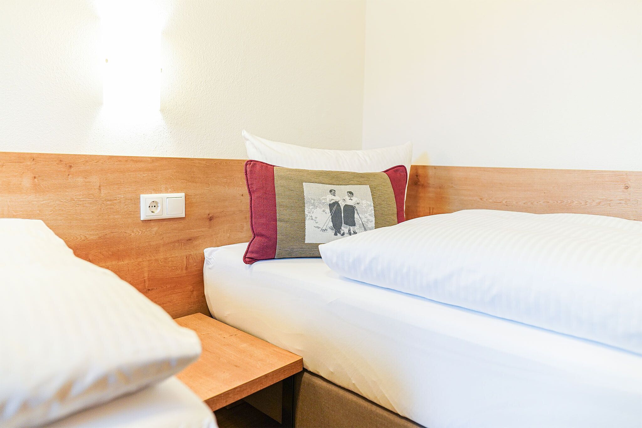 A hotel room with a wooden headboard, a single bed with white bedding, a bedside table, and a wall socket.