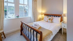 Une chambre avec un lit en bois, du linge de lit blanc, une fenêtre avec des rideaux, une table de chevet avec une lampe et une vue sur l’extérieur.