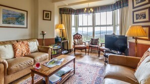 Una sala de estar con sofá, sillón, mesa de centro y televisor. Se observan libros, una lámpara y una ventana con vistas.