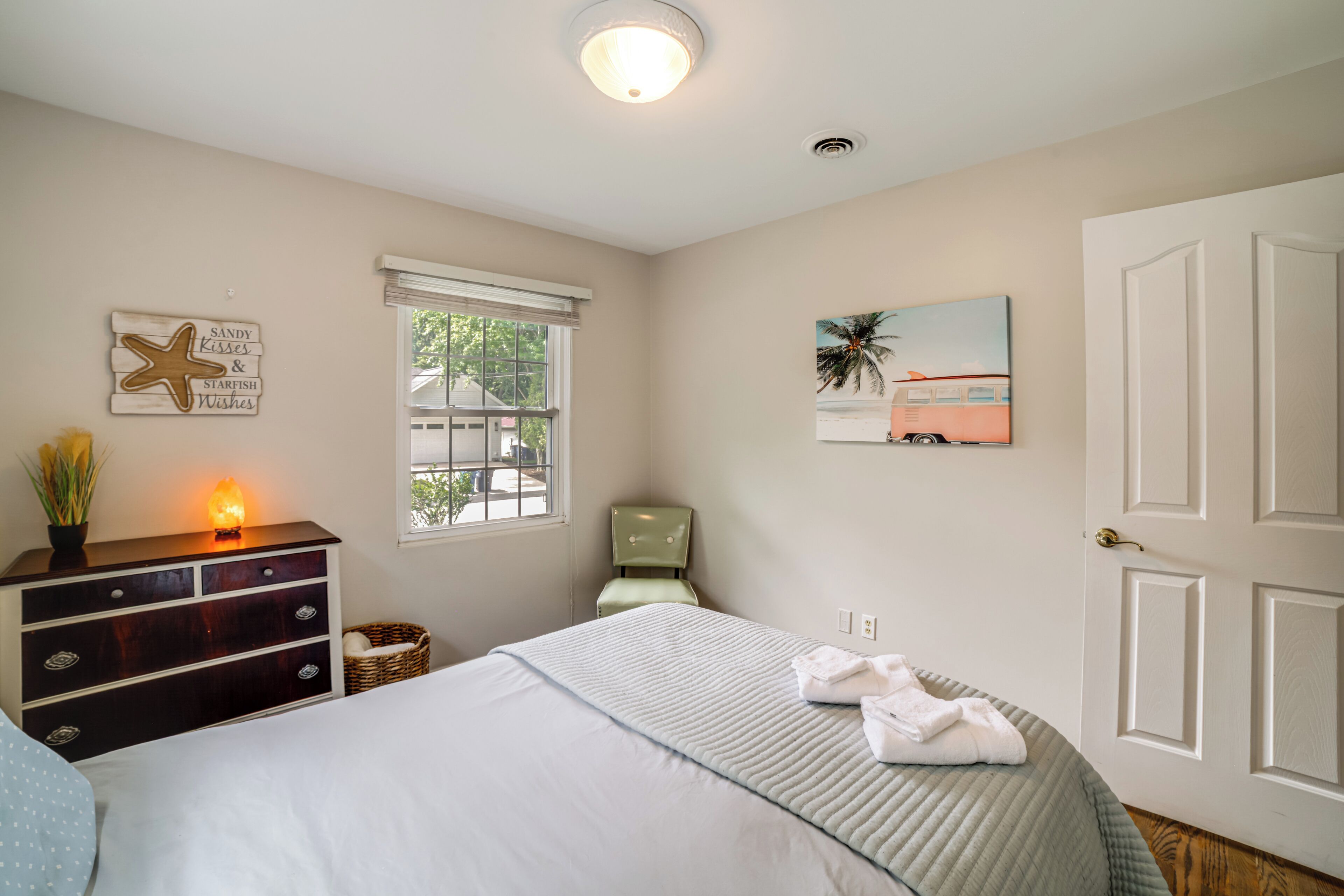 Une chambre à coucher comprenant un lit, une commode, une chaise, une fenêtre et des œuvres d’art.