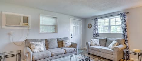 Una sala de estar con dos sofás, una mesa de centro de vidrio, una puerta blanca y una ventana con cortinas estampadas.