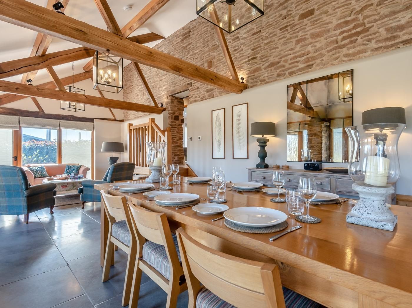 A dining area with a wooden table set for six, flanked by chairs, in a room with exposed wooden beams and a stone wall.
