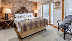 Une chambre avec un mur en pierre, un lit recouvert d’une courtepointe à carreaux, deux lampes de chevet et une chaise près de la fenêtre.