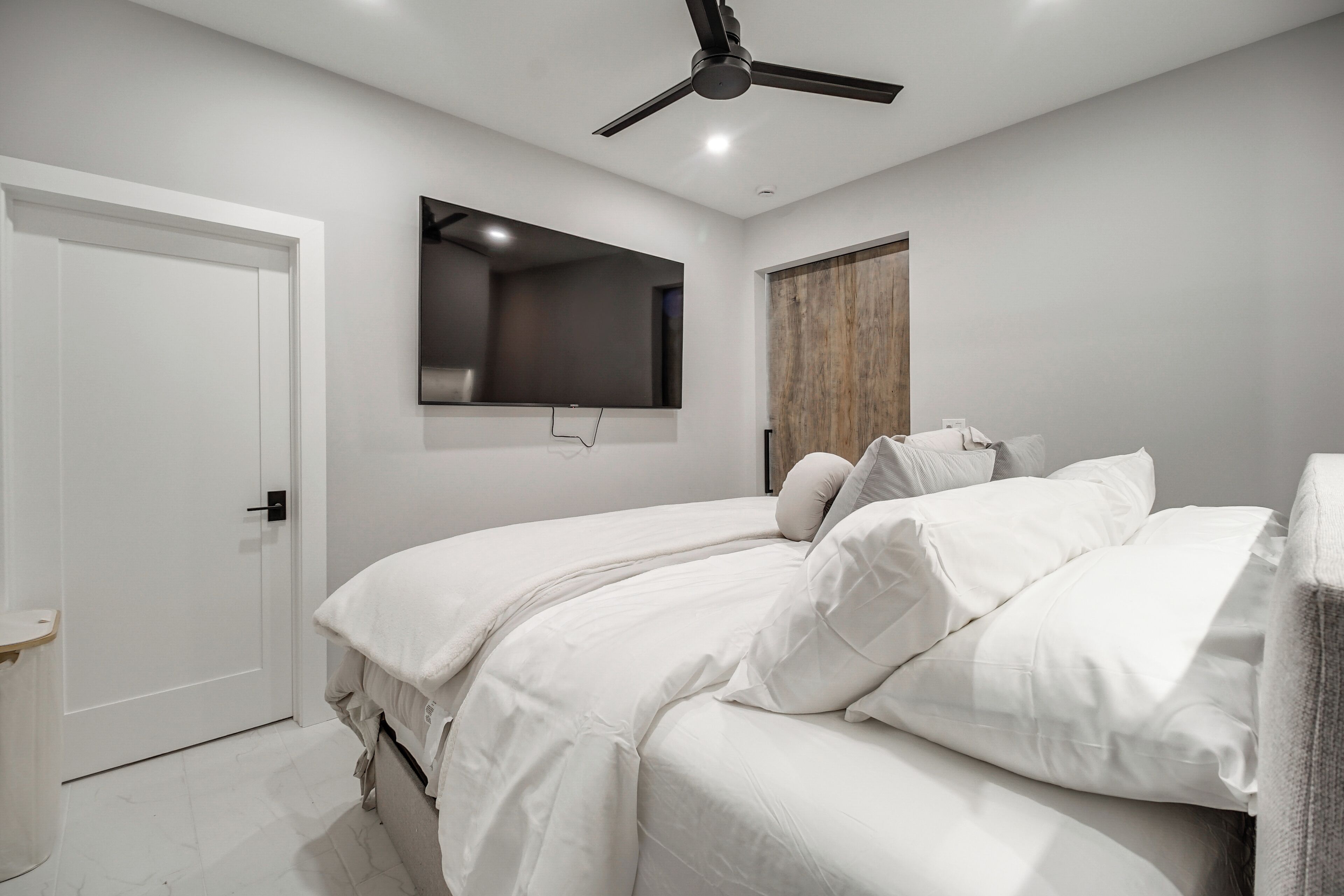 Un dormitorio con cama, ventilador de techo, televisor montado y puerta de madera.