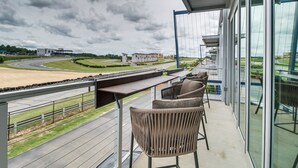 A balcony with a view of a racetrack, outdoor seating, and a modern building with large windows. - Trackside Luxury at Atlanta Motorsports Park (Dawsonville)