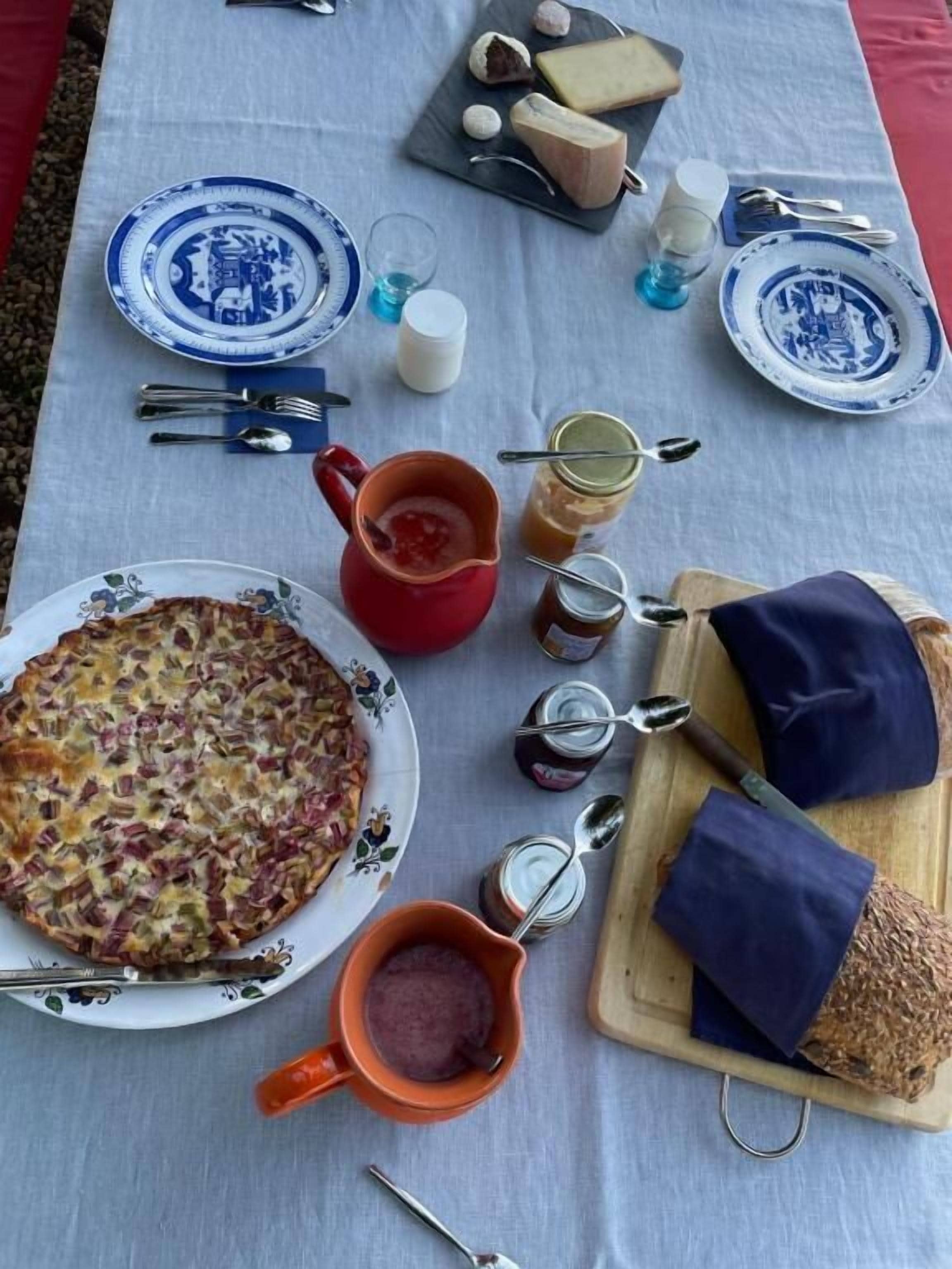A table set with plates, cutlery, a red mug, jars, a wooden board with bread, and a cheese board.