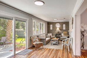 A spacious living room with a large sliding glass door leading to a patio, a wooden floor, and a mix of wicker and upholstered furniture. - Palm Cottage: Walk to Darien Waterfront & Fort (Darien)