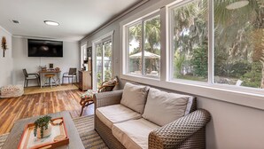 Una sala de estar con sofá, televisión, comedor y ventanas amplias con vista a árboles.