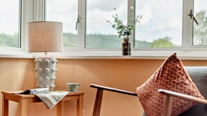 Una habitación acogedora con una mesita de madera, una lámpara, un sillón con un cojín estampado y un florero con flores en un alféizar.