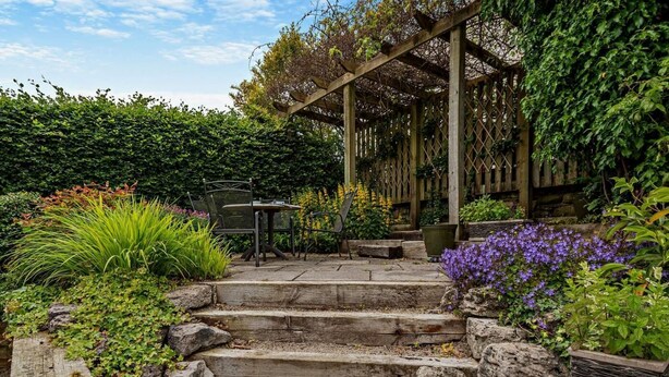 Un jardín con escalones de piedra que conducen a un porche de madera, rodeado de diversas plantas y una mesa con sillas.