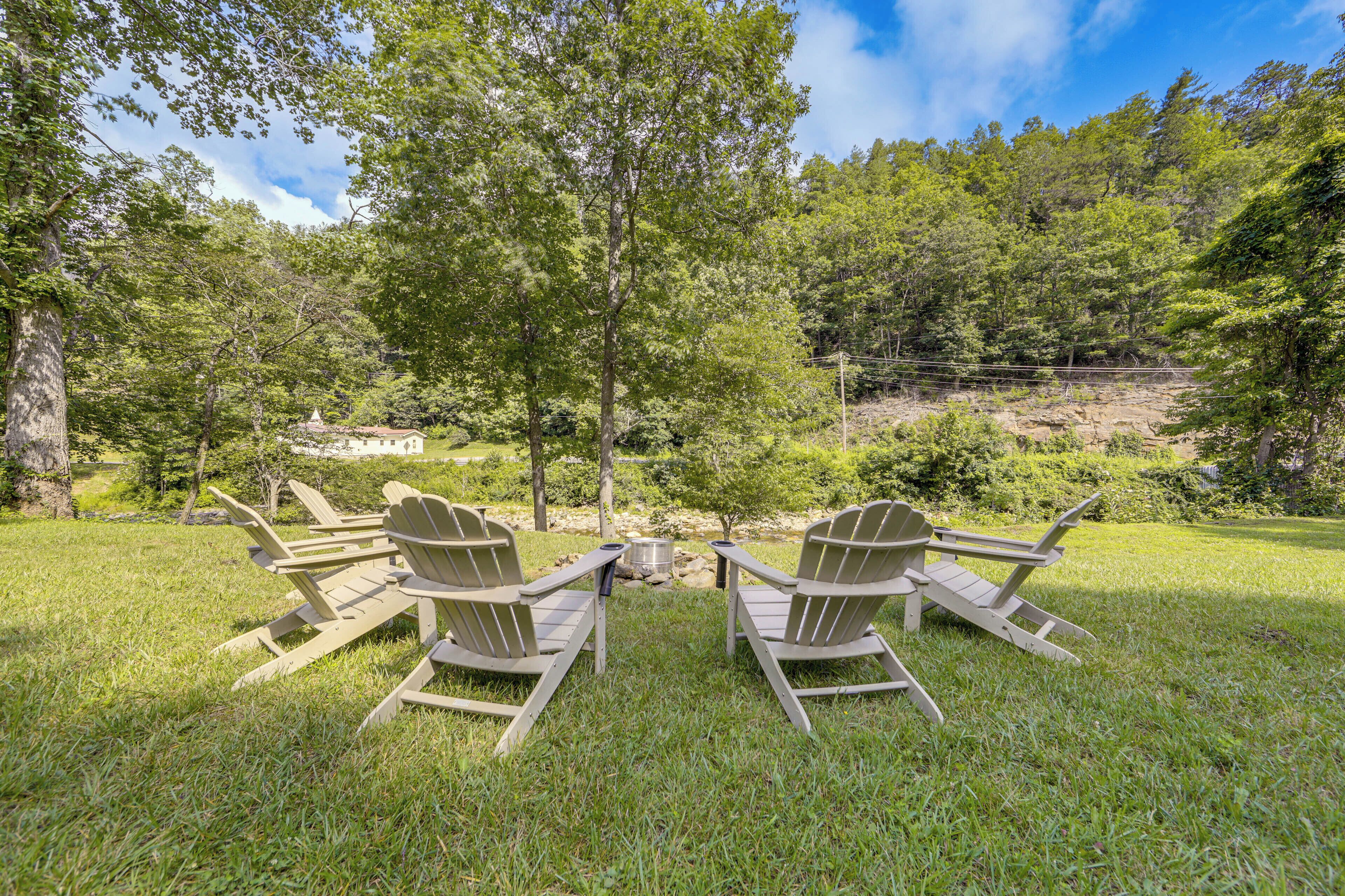 A grassy outdoor area with Adirondack chairs arranged in a semi-circle, a small fire pit, and a backdrop of trees and hills.