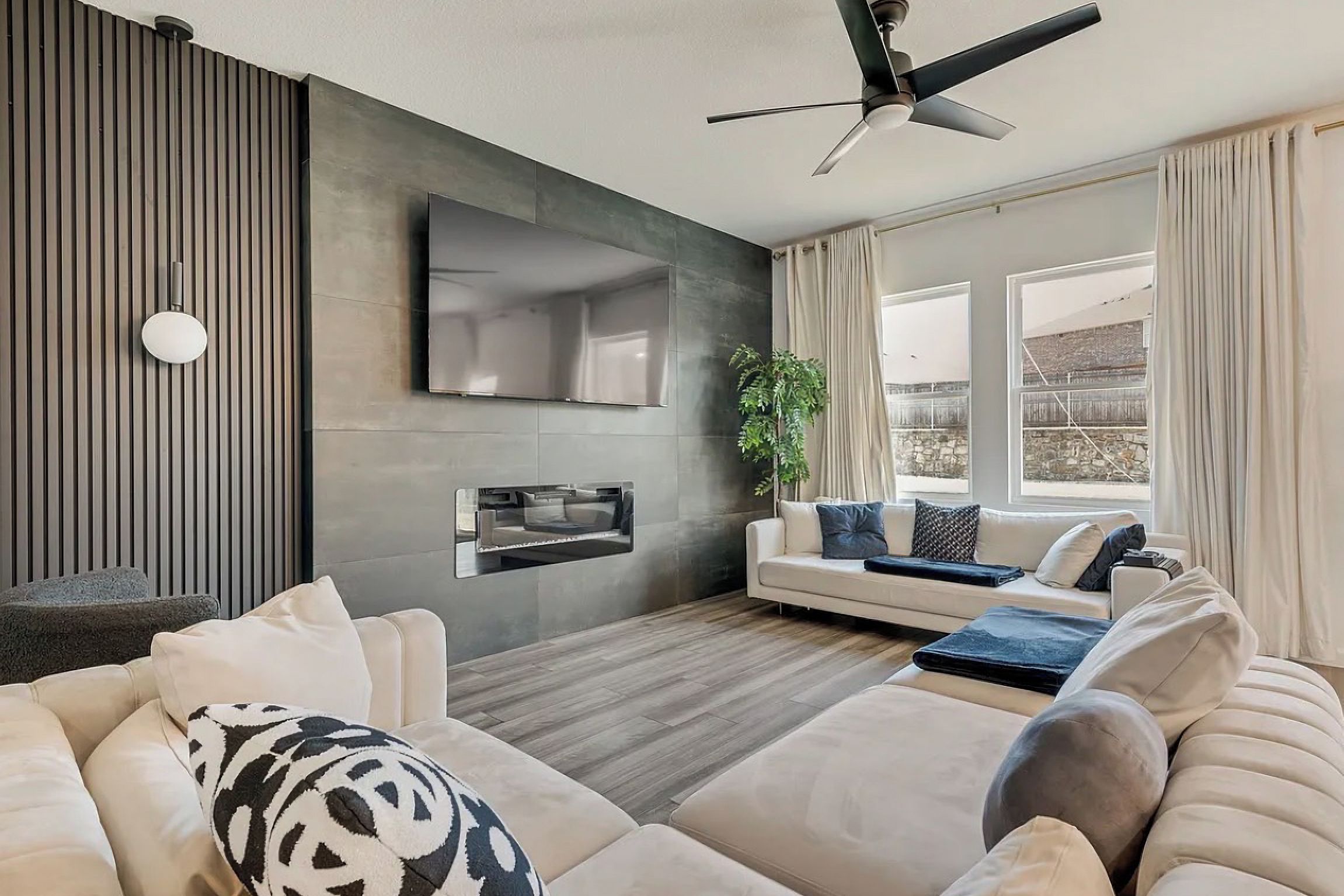 Een moderne woonkamer met een bank, een flatscreen-tv, een plafondventilator en een groot raam met gordijnen.
