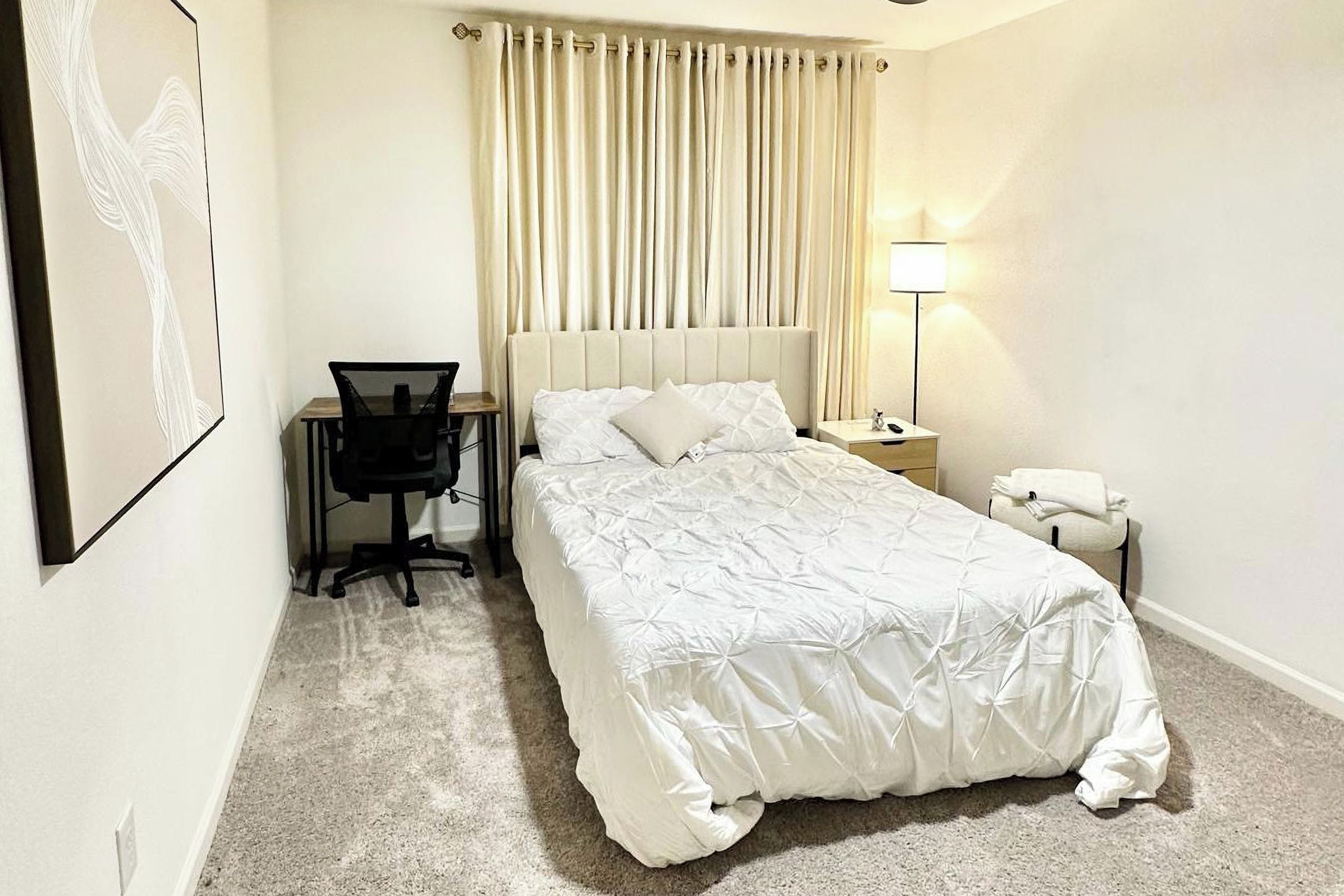 Una habitación de hotel con una cama, un escritorio, una silla, una mesita de noche, una lámpara y una cortina.