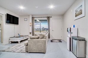 A modern living room with a sofa, a flat-screen TV, a small table, a refrigerator, and a window with curtains. - Family Fun! Quaint Seminole Home w/ Grill & Patio (Seminole)