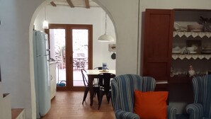 Uma sala de estar com um sofá, uma área de jantar com cadeiras e uma mesa, uma cozinha com frigorífico, e uma porta de madeira que dá para o exterior.