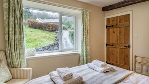 Um quarto com cama, cadeira, janela com cortinas e porta de madeira.