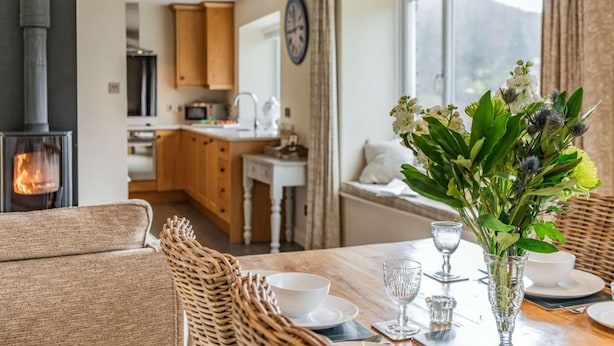 Uma sala de estar aconchegante com lareira, uma mesa de jantar posta com copos e pratos, e vista para a cozinha e o exterior.
