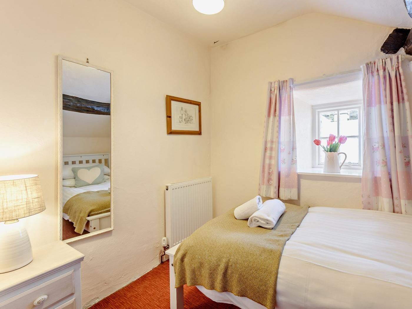 A bedroom with a bed, a bedside table, a lamp, a radiator, a window with curtains, and a framed picture on the wall.