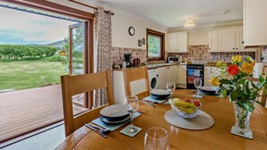 A dining table set with plates, cutlery, and wine glasses, a washing machine, a microwave, and cabinets in a kitchen with a view of a vineyard. - 3 Bed in Dunkeld (oc-50619) (Amulree)