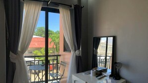 Una habitación con balcón, escritorio, silla y ventana con cortinas.