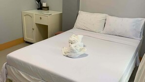Una cama bien hecha con una colcha blanca y almohadas, una mesita de noche pequeña con una lámpara y una taza, y una pared lisa con una puerta.