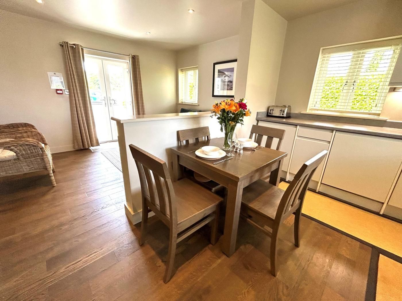 A dining area with a wooden table set for four, a vase with flowers, and a kitchen in the background.