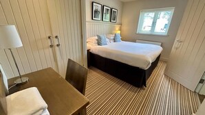 Et hotelværelse med en seng, et skrivebord med stol, en lampe, et vindue med persienner og en dør.
