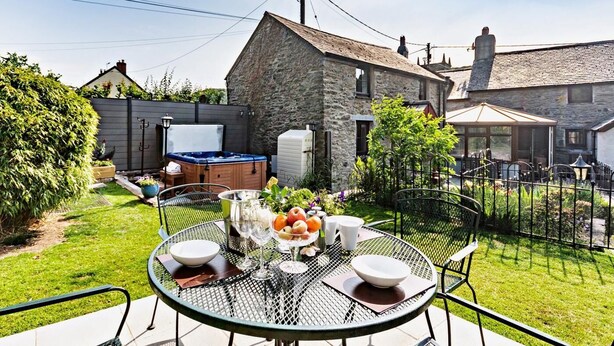 Une maison en pierre avec un jacuzzi, une table ronde en métal entourée de chaises et un jardin clôturé.